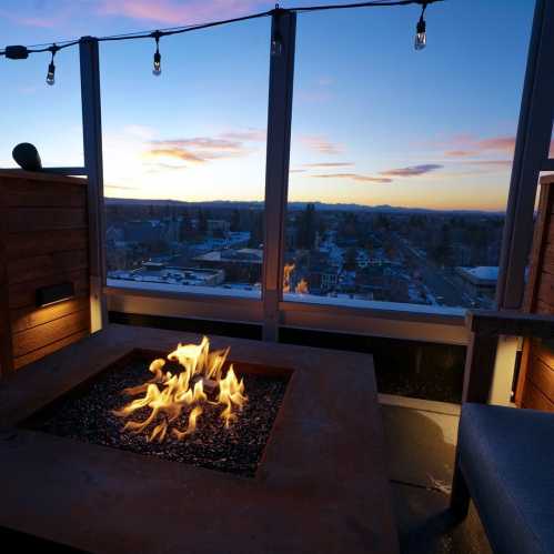 A cozy rooftop setting with a fire pit, overlooking a city skyline at sunset, with string lights above.