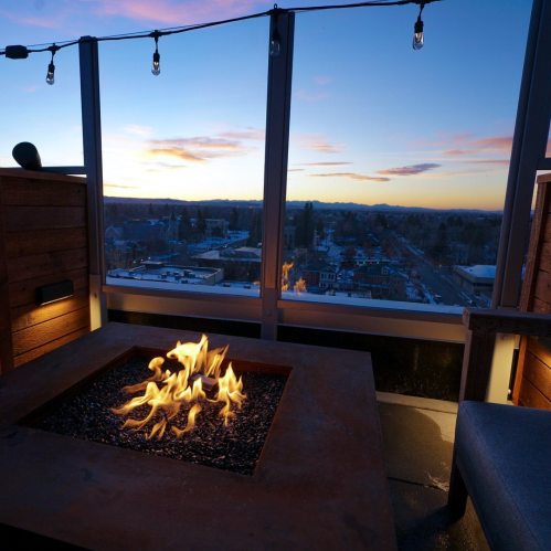 A cozy rooftop setting with a fire pit, overlooking a city skyline at sunset, with string lights above.