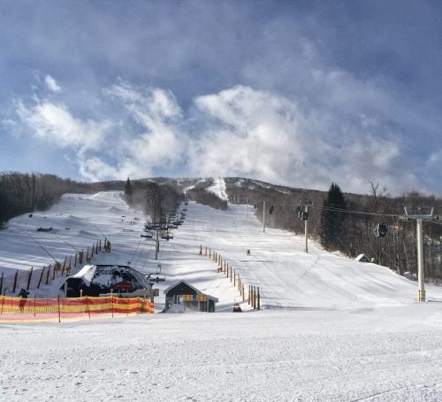 A snowy ski slope with a chairlift, surrounded by trees and a cloudy sky. A small lodge is visible at the base.