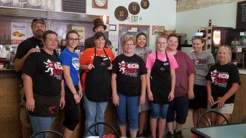 A group of smiling restaurant staff members in black aprons pose together in a cozy diner setting.