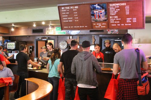 A busy diner scene with customers ordering at the counter and staff preparing food, featuring a menu board overhead.
