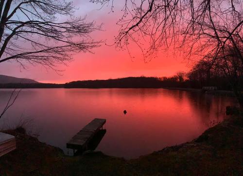 A serene lake at sunset, reflecting vibrant pink and orange hues, with a wooden dock and bare trees in the foreground.