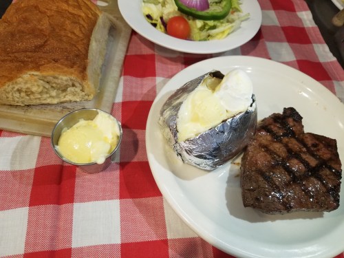 A plate with grilled steak, a baked potato with sour cream, a side of butter, and a loaf of bread on a checkered tablecloth.