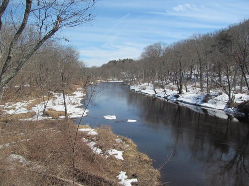 A serene river flows through a winter landscape, with patches of snow and bare trees under a blue sky.