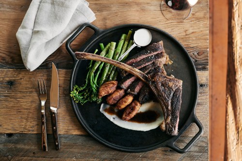 A beautifully plated meal featuring a large steak, asparagus, mashed potatoes, and a small dish of sauce on a wooden table.