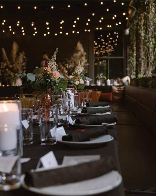 Elegant dining setup with dark tablecloth, floral centerpieces, and soft string lights in a cozy atmosphere.