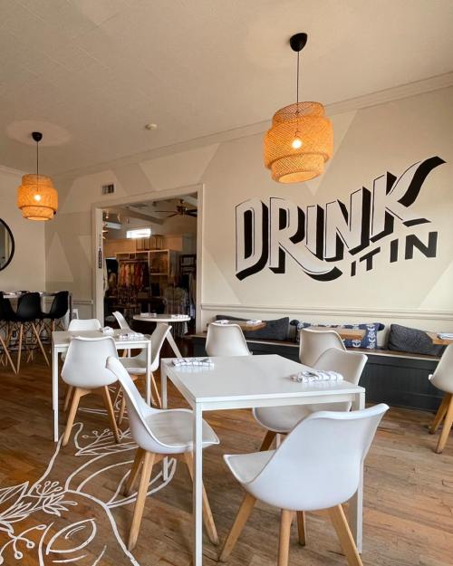 A cozy café interior featuring white tables, modern chairs, and a large "DRINK IT IN" mural on the wall.