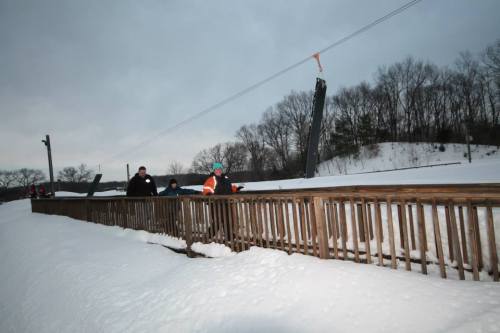 A snowy landscape with a wooden bridge and people walking, surrounded by trees and a cloudy sky.