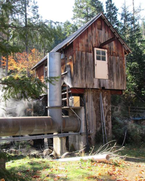 A rustic wooden building on stilts, with a large metal pipe and surrounded by trees and autumn foliage.