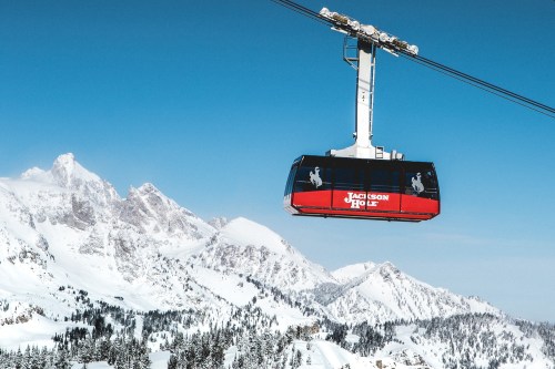A ski gondola suspended above snowy mountains under a clear blue sky.