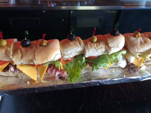 A long sandwich with layers of meats, cheese, lettuce, and tomatoes, topped with olives and pickles on toothpicks.
