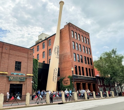 A large baseball bat leans against a brick building, with a crowd of people gathered outside.