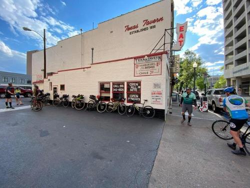 Bicyclists gather outside Texas Tavern, a white building with a sign, in a lively urban setting.