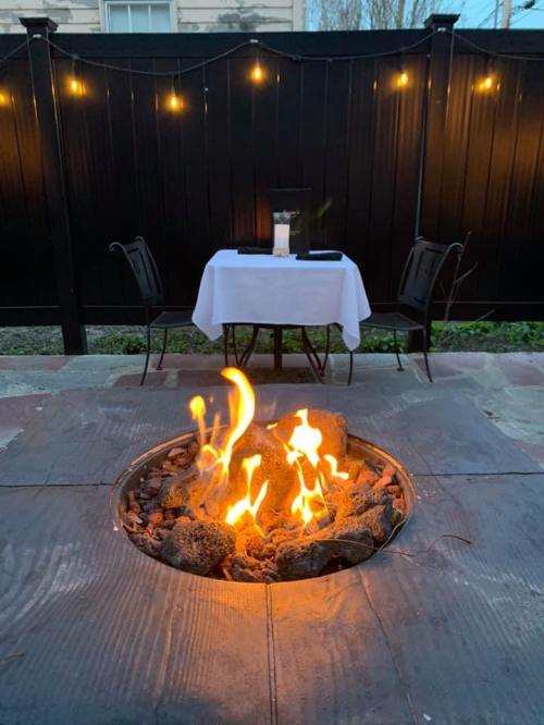 A cozy outdoor dining setup with a table and chairs near a fire pit, illuminated by string lights.
