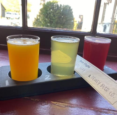 Three small glasses of beer in a flight, featuring orange, pale yellow, and red drinks on a wooden tray.