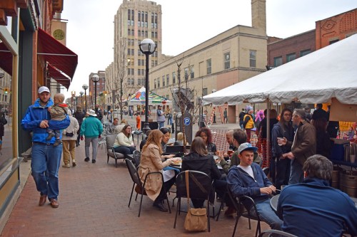 A bustling street scene with people dining outdoors, shopping, and enjoying a festive atmosphere under a cloudy sky.