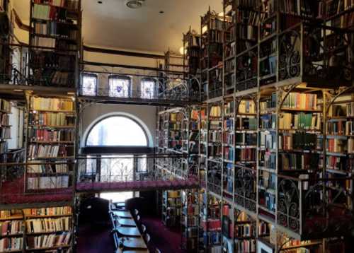 A grand library with multiple levels of bookshelves, ornate railings, and large windows letting in natural light.