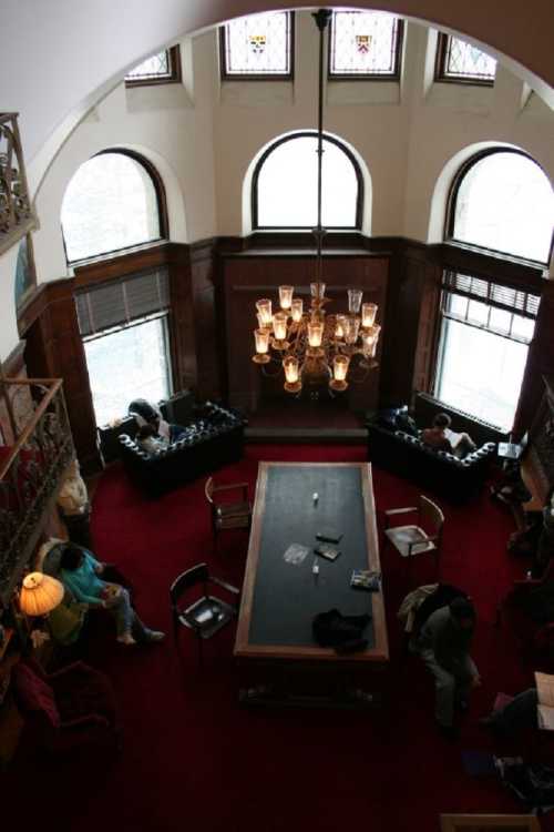 A spacious room with a chandelier, large table, and seating areas, featuring large windows and a red carpet.