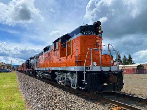 An orange and black locomotive parked on tracks under a cloudy sky, with additional train cars visible behind it.