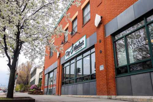 A brick building with a sign reading "City Market," surrounded by blooming trees and a sidewalk.