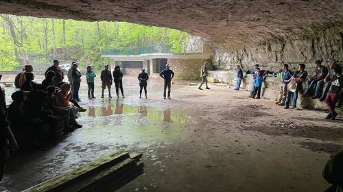 A group of people gathered in a cave-like space, some sitting and others standing, with greenery visible outside.
