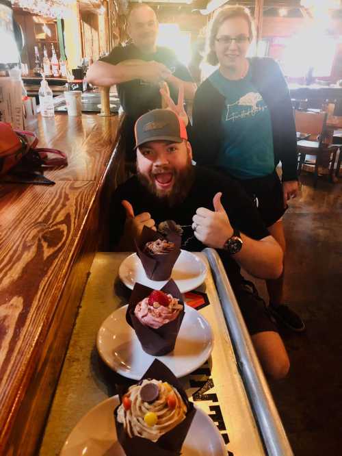 A man with a beard gives a thumbs up in front of dessert cups, while two people smile in the background at a restaurant.