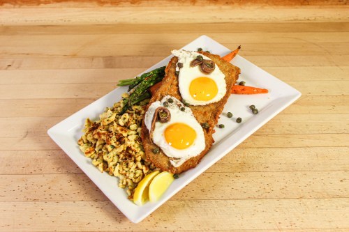 A plate of fried eggs on breaded meat, served with asparagus, carrots, and a side of scrambled eggs with capers.