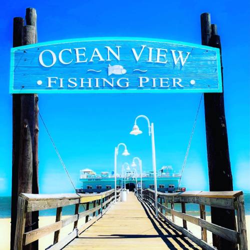 A wooden fishing pier with a blue sign reading "Ocean View Fishing Pier" under a clear blue sky.
