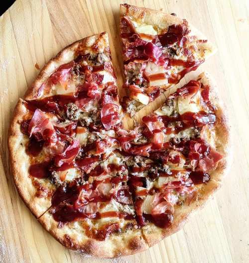 A round pizza topped with prosciutto, pineapple, and barbecue sauce, with one slice removed.