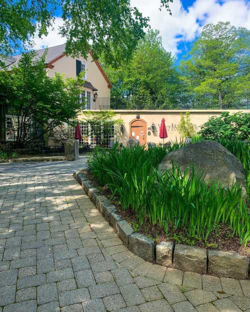 A stone pathway leads to a charming building surrounded by lush greenery and a large rock in the garden.