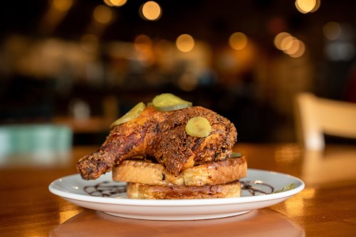A plate featuring a crispy fried chicken leg on toasted bread, topped with pickles, set in a warm, blurred restaurant background.