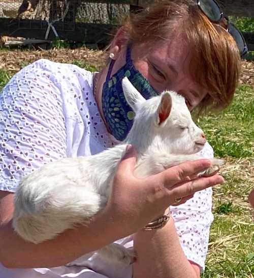 A woman in a mask gently holds a small white goat in her hands, smiling in a sunny outdoor setting.