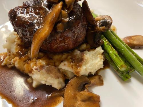 A plated dish featuring a beef filet topped with mushrooms, served on mashed potatoes with asparagus and a rich sauce.