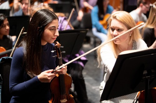 Two musicians in an orchestra practice together, one holding a violin and the other guiding with a bow.