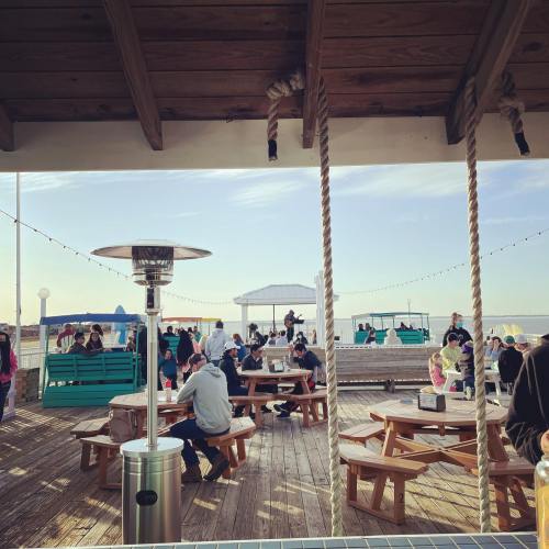 A lively outdoor space with people sitting at picnic tables, enjoying the sun near the water.