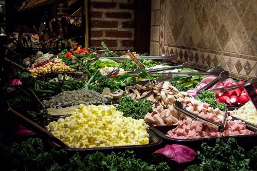 A colorful buffet display featuring various fresh vegetables, meats, and toppings in serving trays.