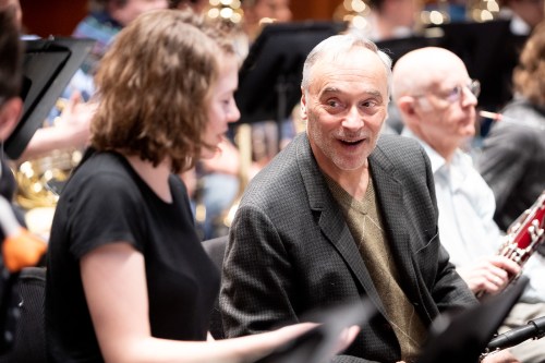 A man in a blazer smiles while talking to a woman in a black shirt during a music rehearsal with other musicians in the background.