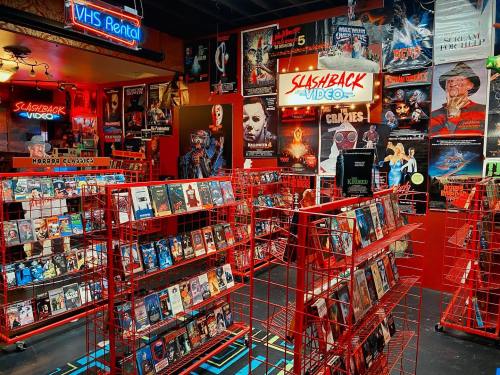 A vibrant video rental store filled with red racks of VHS tapes and horror movie posters on the walls.