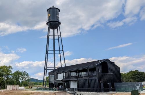 A black water tower stands beside a modern building against a blue sky with clouds and green hills in the background.