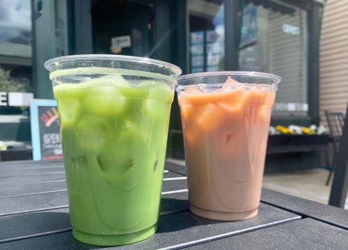 Two clear cups filled with green and brown iced drinks sit on a table outside a café.