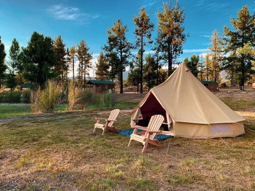 A tan tent set up in a grassy area with two wooden chairs, surrounded by trees and a clear blue sky.