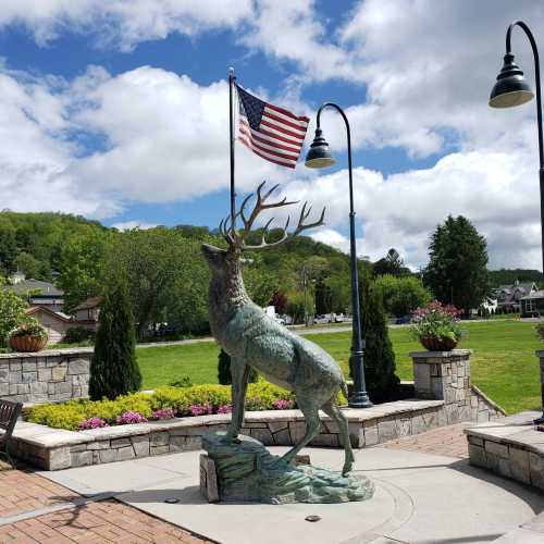 A bronze deer statue stands near a flagpole with an American flag, surrounded by greenery and flowers.