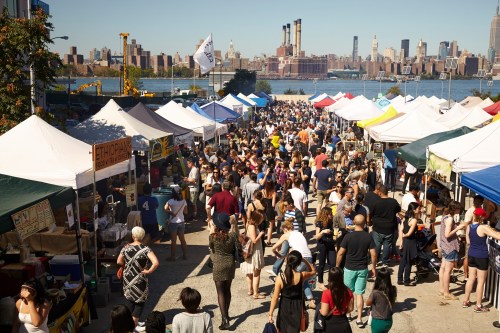A bustling outdoor market with colorful tents, crowds of people, and a city skyline in the background by the water.