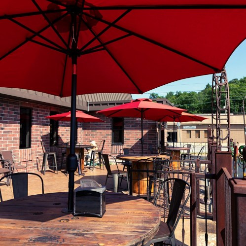 Outdoor seating area with red umbrellas, wooden tables, and a brick building in the background.