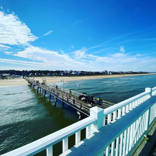 A wooden pier extends over calm water, with a sandy beach and distant buildings under a clear blue sky.