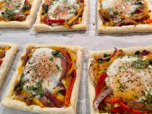 Baked pastry squares topped with eggs, colorful bell peppers, and herbs on a parchment-lined tray.