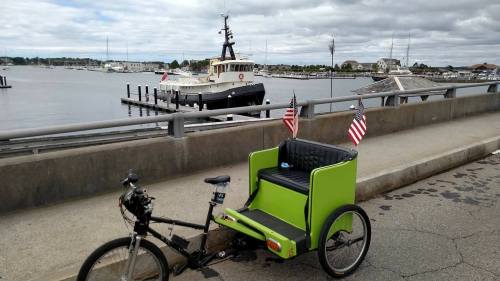 A green rickshaw with American flags parked by a harbor, with boats and a cloudy sky in the background.