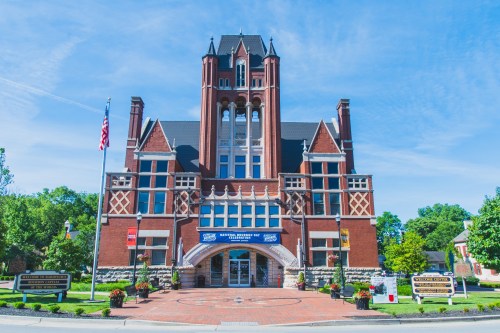 Historic brick building with a tall tower, blue sky, and American flag, featuring banners and landscaped surroundings.