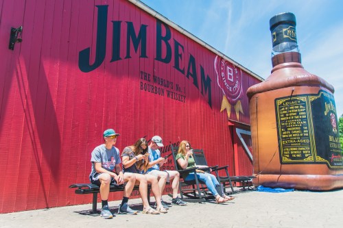 Four people sit on a bench in front of a large Jim Beam whiskey bottle mural on a bright, sunny day.