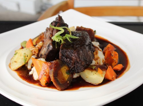 A plate of tender beef served over mashed potatoes, garnished with vegetables and a rich brown sauce.